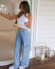 Woman wearing a white tank top and blue jeans standing in a room with wooden flooring.
